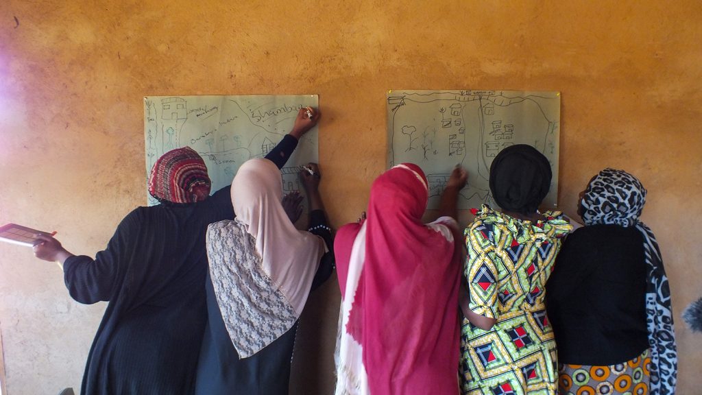 Women at a pale orange wall with 2 posters stuck to it, which women are adding to.